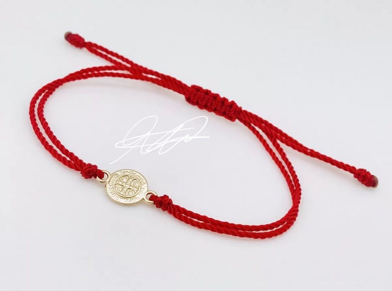 May include: Red string bracelet with a gold-colored medallion. The medallion features a detailed design. The bracelet is adjustable with a sliding knot closure. The design is simple and elegant.