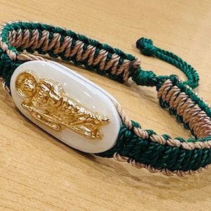 May include: A green and brown braided bracelet with a white oval charm featuring a gold image of a saint.