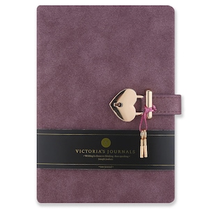 Könnte beinhalten: Ein violettes Journal mit Wildlederoptik und einem goldfarbenen herzförmigen Vorhängeschloss, an dem zwei kleine Schlüssel mit einer rosa Schnur befestigt sind. Ein schwarzes Papierband umschließt die Mitte mit dem Text Victoria's Journals.