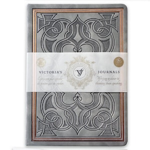 Puede incluir: Un diario rectangular con una cubierta gris con un diseño en relieve y ornamentado. El diario tiene un borde color cobre y una banda blanca con el texto "VICTORIA'S JOURNALS". La cubierta del diario tiene el texto "Writing is closer to thinking than speaking".