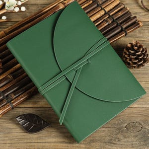 May include: A rectangular dark green journal with a smooth, solid-colored cover. The journal features a large, semi-circular flap that folds over the front, secured by a thin, matching green strap that wraps around the cover in a crisscross pattern.
