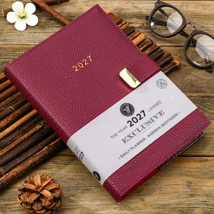 May include: A deep magenta 2027 daily planner with a textured cover, a gold-toned metal clasp, and a white paper band featuring the text: THE YEAR 2027 EXCLUSIVE L'ANNÉE DAILY PLANNER AGENDA QUOTIDIEN.