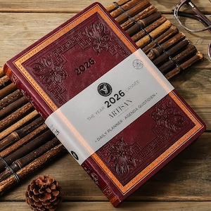 Può includere: Un'agenda giornaliera Artisan 2026 bordeaux con disegni floreali e geometrici in rilievo. L'agenda ha finiture color oro e una fascia bianca con il testo "The Year 2026 L'Année Artisan Daily Planner Agenda Quotidien."
