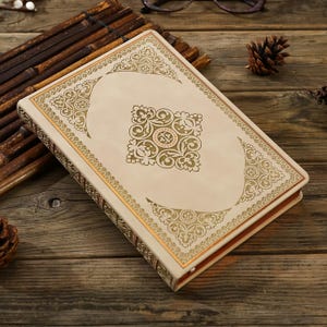 May include: Cream-coloured journal with ornate gold filigree design on the cover and spine. The cover features a central square design and decorative borders. The journal is lying on a wooden surface with a bamboo mat and pine cones.