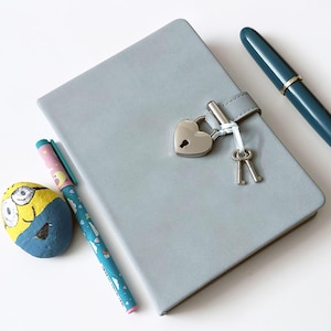 Könnte beinhalten: Ein hellgraues Notizbuch mit herzförmigem Schloss und zwei Schlüsseln. Ein blauer Stift und ein bunter Stift liegen neben dem Notizbuch. Ein bemalter Stein mit einer Cartoonfigur ist ebenfalls im Bild.