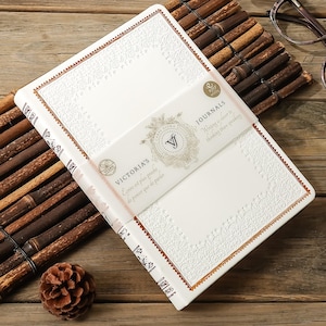 Peut inclure: Un journal blanc avec des motifs floraux en relief et une bordure décorative. Le journal est enveloppé d'une bande qui indique "VICTORIA'S JOURNALS" et comprend le texte "Writing is closer to thinking than speaking." Un cône de pin et un tapis en bambou sont en arrière-plan.