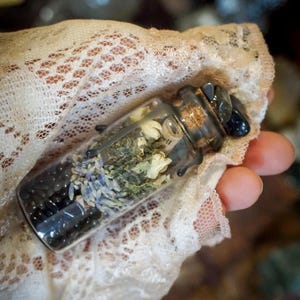May include: A small glass bottle filled with dried herbs, flowers, and black beads. The bottle has a cork stopper and is tied with a black ribbon.