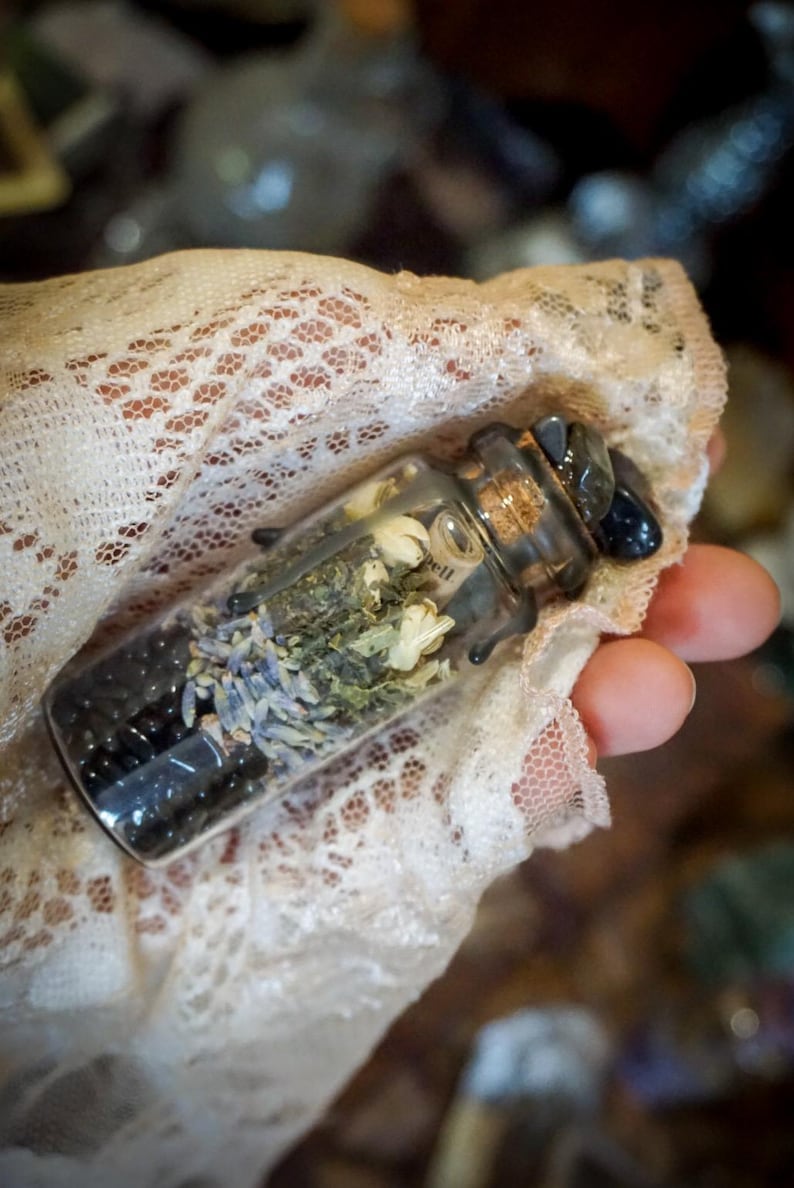 May include: A small glass bottle filled with dried herbs, flowers, and black beads. The bottle has a cork stopper and is tied with a black ribbon.