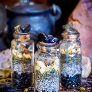 May include: Three small glass bottles filled with various herbs, crystals, and dried flowers. The bottles are sealed with cork stoppers and have a black wax seal. The bottles are arranged on a colorful patterned fabric.