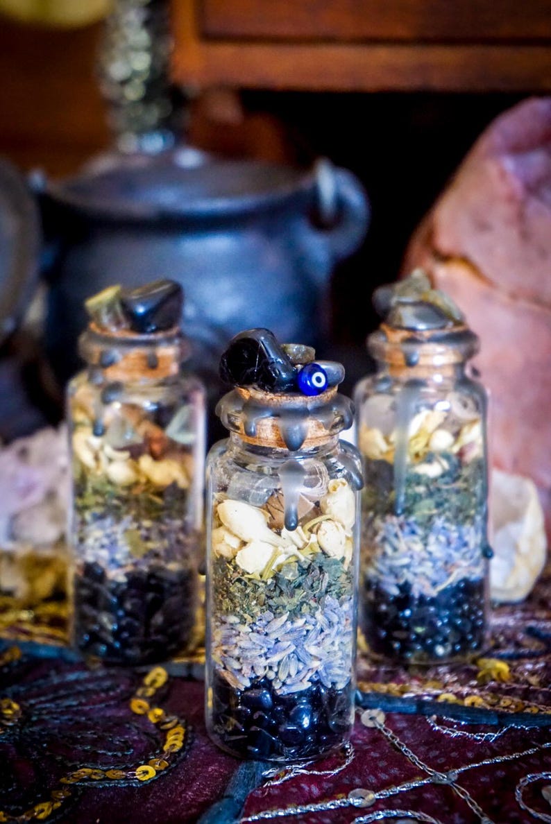 May include: Three small glass bottles filled with various herbs, crystals, and dried flowers. The bottles are sealed with cork stoppers and have a black wax seal. The bottles are arranged on a colorful patterned fabric.