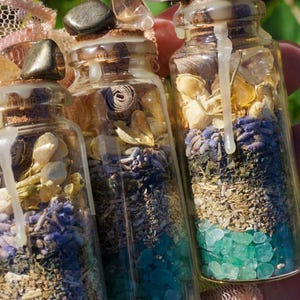 May include: Three small glass bottles filled with various dried herbs, flowers, and crystals. The bottles are topped with cork stoppers and sealed with wax. The bottles are arranged in a row, with the largest bottle in the back and the smallest bottle in the front.