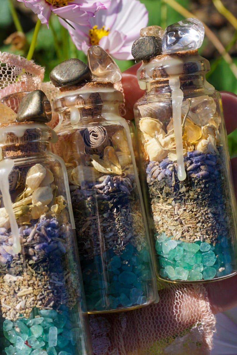 May include: Three small glass bottles filled with various dried herbs, flowers, and crystals. The bottles are topped with cork stoppers and sealed with wax. The bottles are arranged in a row, with the largest bottle in the back and the smallest bottle in the front.