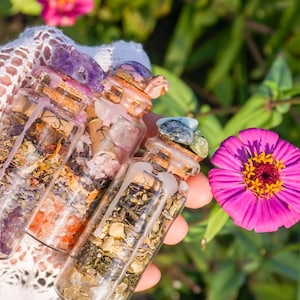 May include: Three small glass bottles filled with various herbs, crystals, and dried flowers. The bottles have cork stoppers and are sealed with wax. The bottles are held in a hand against a green and pink floral background.