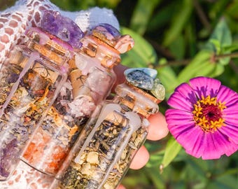 May include: Three small glass bottles filled with various herbs, crystals, and dried flowers. The bottles have cork stoppers and are sealed with wax. The bottles are held in a hand against a green and pink floral background.