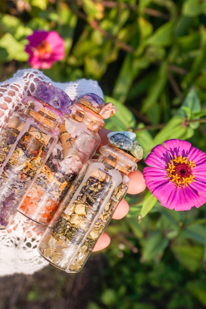 May include: Three small glass bottles filled with various herbs, crystals, and dried flowers. The bottles have cork stoppers and are sealed with wax. The bottles are held in a hand against a green and pink floral background.