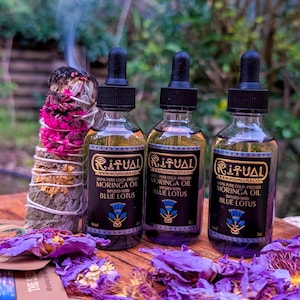 May include: Three glass bottles of Ritual Moringa Oil infused with Blue Lotus, each with a black dropper. The bottles are on a wooden surface, surrounded by purple flower petals and a bundle of burning sage. The bottles have a black label with gold text.