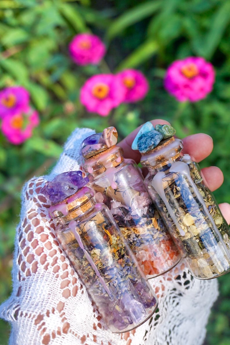 May include: Three small glass bottles filled with dried herbs and crystals. The bottles are topped with cork stoppers and sealed with wax. The bottles are held in a hand wearing a white lace glove.