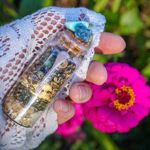 May include: A small glass bottle filled with various herbs, crystals, and a turquoise stone. The bottle is sealed with a cork and wax. The bottle is held in a hand wearing a white lace glove.