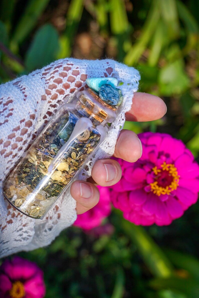 May include: A small glass bottle filled with various herbs, crystals, and a turquoise stone. The bottle is sealed with a cork and wax. The bottle is held in a hand wearing a white lace glove.