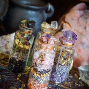 May include: Four small glass bottles filled with various crystals, herbs, and other natural elements. The bottles are topped with cork stoppers and sealed with wax. The bottles are arranged on a patterned fabric surface.