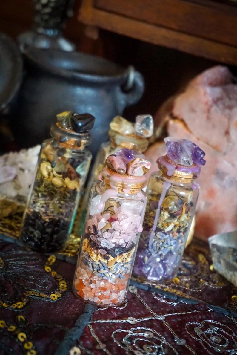 May include: Four small glass bottles filled with various crystals, herbs, and other natural elements. The bottles are topped with cork stoppers and sealed with wax. The bottles are arranged on a patterned fabric surface.