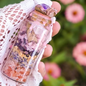 May include: A small glass bottle filled with pink and purple crystals, dried flowers, and a rolled up piece of paper. The bottle is topped with a cork and a purple amethyst stone.