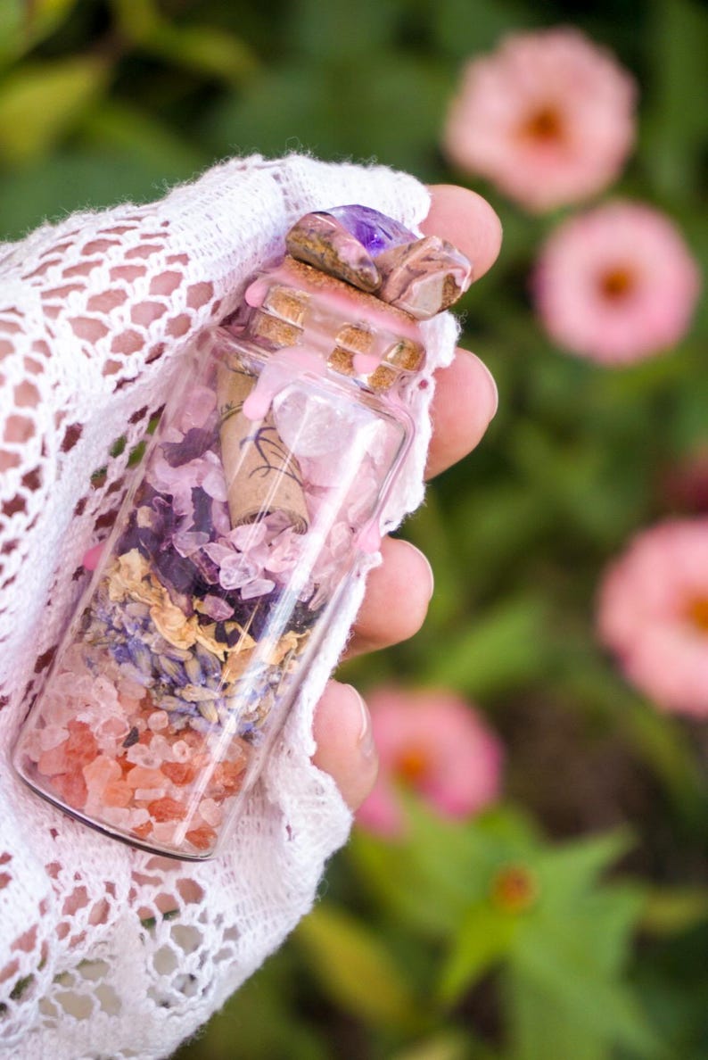 May include: A small glass bottle filled with pink and purple crystals, dried flowers, and a rolled up piece of paper. The bottle is topped with a cork and a purple amethyst stone.