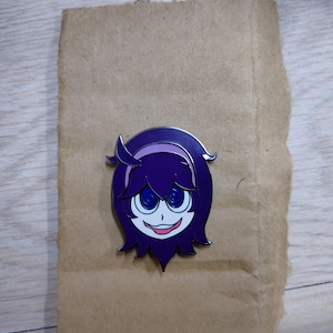 Puede incluir: Pin de esmalte morado y rosa con un personaje de dibujos animados con ojos en espiral y una amplia sonrisa.