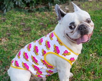 May include: A white French bulldog wearing a white tank top with a pink heart and yellow "LOVE" print. The dog is sitting on green grass.