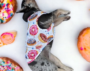 May include: A French bulldog wearing a blue and white tank top with a donut print. The dog is lying on a white surface surrounded by donuts with colorful sprinkles.