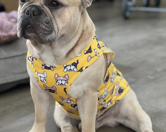 May include: A French Bulldog wearing a yellow tank top with a pattern of cartoon French Bulldogs. The tank top has a black strap.