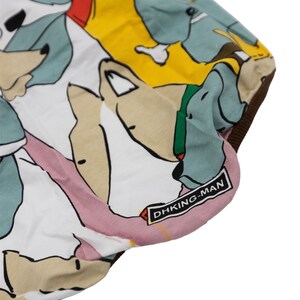 May include: White fabric with a colorful print of cartoon dogs. The fabric has a pink, yellow, and blue background with brown, black, and white dogs. The fabric has a brown border and a black label that says "DHKING-MAN".