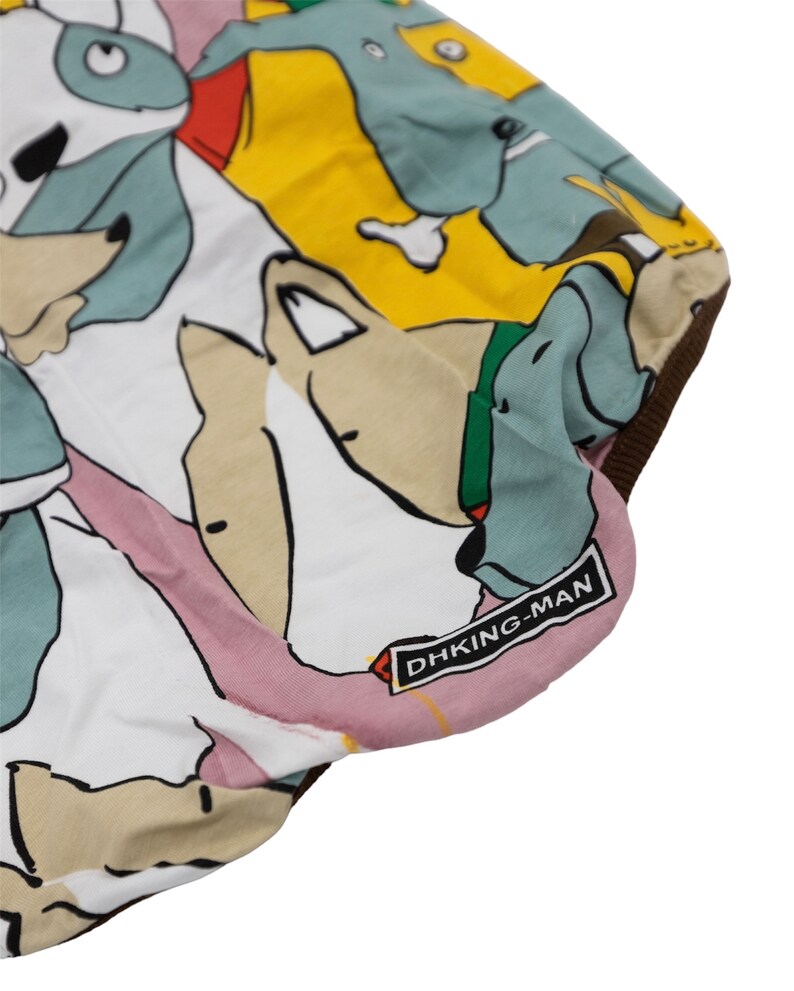 May include: White fabric with a colorful print of cartoon dogs. The fabric has a pink, yellow, and blue background with brown, black, and white dogs. The fabric has a brown border and a black label that says "DHKING-MAN".