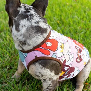 May include: A French bulldog wearing a pink and brown dog tank top with a colorful cartoon print. The dog is sitting on green grass.