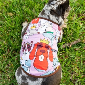 May include: A French bulldog wearing a pink dog shirt with a multi-colored print of cartoon dogs wearing crowns. The shirt has a brown trim around the neck and armholes.