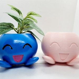 May include: Two whimsical, round planters with smiling faces. One is blue with a pink tongue and holds a small plant. The other is pink. Both have small, rounded feet. These decorative planters are perfect for small plants.