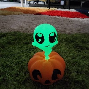 May include: A green alien figurine with large eyes sits atop an orange pumpkin with a carved face. The pumpkin is on a green shag carpet.