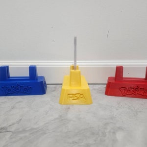 May include: Three Pokemon card display stands, one blue, one yellow, and one red. The yellow stand has the text "PSA" on the front. All three stands have the Pokemon logo on the front.