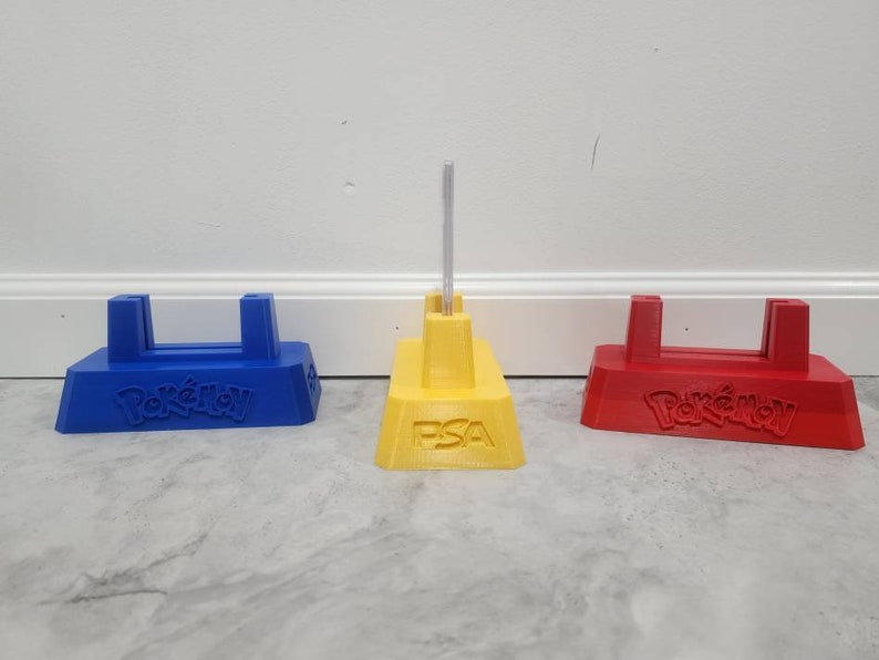 May include: Three Pokemon card display stands, one blue, one yellow, and one red. The yellow stand has the text "PSA" on the front. All three stands have the Pokemon logo on the front.