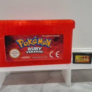 May include: A red Game Boy Advance game cartridge case with the text "Pokémon Ruby Version" and the Nintendo logo. The case is sitting on a white shelf next to a black Game Boy Advance game cartridge.