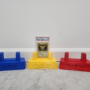 May include: Three Pokemon card display stands, one blue, one yellow, and one red. Each stand has the word "Pokemon" printed on the front. A holographic Pokemon card is displayed on the yellow stand.