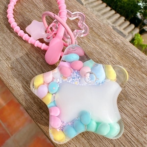 Könnte beinhalten: Ein sternförmiger Schlüsselanhänger mit einem transparenten Harzkörper, gefüllt mit pastellfarbenen Kugeln und Glitzer. Der Schlüsselanhänger hat einen rosa Silikonriemen, einen sternförmigen Anhänger und einen rosa Verschluss.