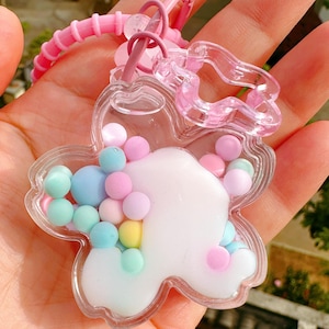 May include: A clear, flower-shaped keychain filled with colorful pastel beads. The keychain has a pink strap and a pink flower-shaped ring. The beads are in shades of blue, pink, yellow, and green. The keychain is held in a hand.