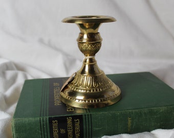 May include: A gold metal candle holder with an ornate design. The holder is sitting on top of a green book titled "Properties of Engineering Materials".