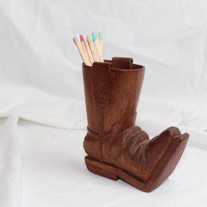 May include: A brown wooden cowboy boot-shaped container holding several matches with colorful tips. The boot has detailed carvings and is set against a white background.