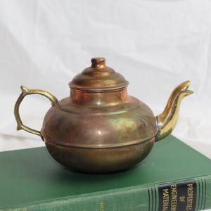 May include: A vintage brass teapot with a rounded body, a lid, and a curved spout. The handle and spout have a golden hue, contrasting with the darker, aged brass of the body. The teapot sits on a green book against a white background.