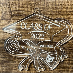 May include: White acrylic ornament shaped like a graduation cap with a scroll and a bulldog mascot. The ornament says "Class of 2022" and "Michael" and "NCA & T".