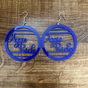 May include: A pair of circular blue earrings featuring the text Aggie Pride cut out from the centre. Each earring is suspended from a silver-toned metal hook. The design consists of a solid blue circular frame with horizontal bars.