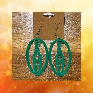 May include: A pair of green oval earrings with the letters "FAMU" cut out. The earrings hang from silver-colored hooks and are displayed on a brown cardboard backing.