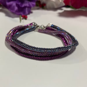 May include: A set of three beaded bracelets in shades of purple and blue. The bracelets are made with small, iridescent beads and have a silver clasp.